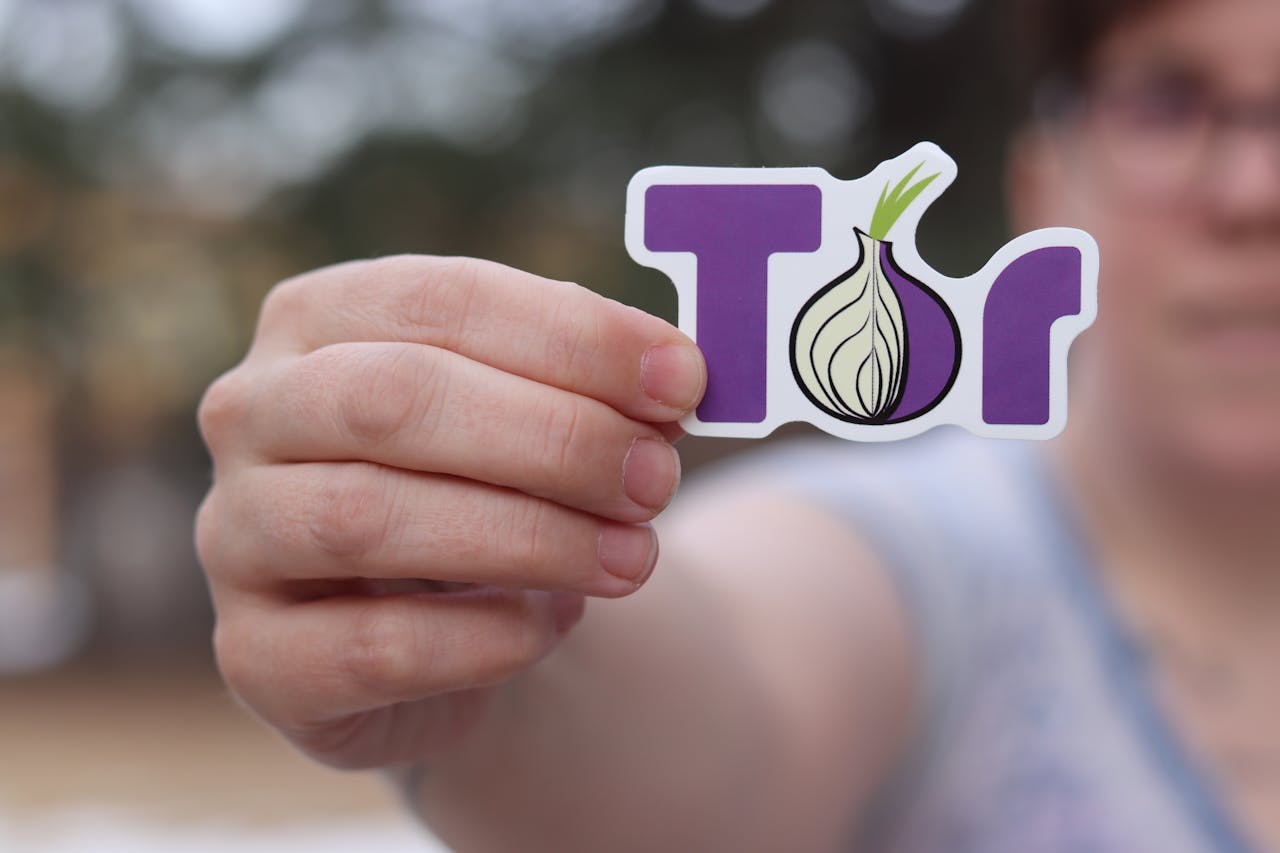 hero-img-02 A close-up of a person holding a Tor browser logo sticker with a blurred outdoor background.